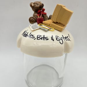 May include: A clear glass jar with a cream-colored lid, decorated with a brown teddy bear, a vintage computer, and the text "Nibbles, Bits & Bytes!". The bear has a red bow. A yellow paperclip and small papers are also on the lid.