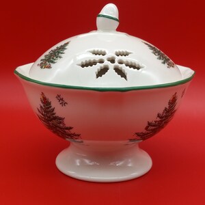 May include: White ceramic Christmas dish with a lid featuring a snowflake-shaped vent. The dish is decorated with Christmas tree designs and a green trim. The dish sits on a pedestal base.