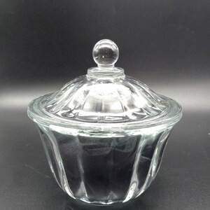 May include: A clear glass candy dish with a lid. The dish has a scalloped design and a round knob on top of the lid. The glass is transparent, allowing a view of the contents inside. The dish is likely used for storing candies or small items.