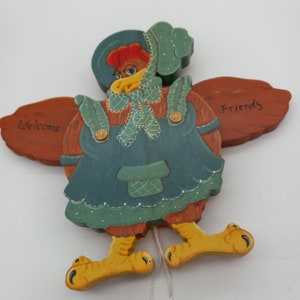 May include: A wooden pull toy shaped like a hen wearing a blue dress, a green apron, and a blue and white hat. The toy has yellow feet and a string attached to its body. The wings are painted with the words "Welcome" and "Friends".