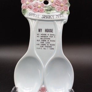 May include: A light blue ceramic spoon rest with two spoon-shaped holders. The top features pink flowers and the text "GREAT SMOKY MTS." Below, the text reads "MY HOUSE" and a poem. The spoon rest is supported by a clear plastic stand.