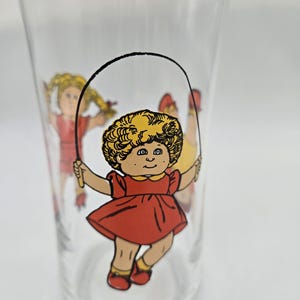 May include: Clear glass tumbler featuring a colorful illustration of a girl with yellow hair in a red dress, jumping rope. Additional figures are visible in the background. The tumbler is a vintage collectible.