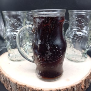 May include: Three glass mugs, two empty and one filled with a dark brown liquid. The mug with the liquid has the word "Frostie" and "Root Beer" printed on it.