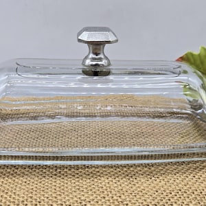 May include: A clear glass butter dish with a rectangular base and a domed lid. The lid has a silver-colored, geometric handle. The dish sits on a burlap surface, with a white and yellow flower in the background.