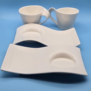 May include: Two white ceramic cups with curved handles paired with two matching rectangular white ceramic saucers. Each saucer features a distinct wave-like design with a circular indentation specifically shaped to hold one of the cups securely.