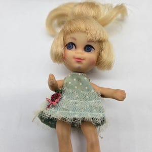 May include: A vintage doll with blonde hair and blue eyes. The doll wears a light green dress with white polka dots and a pink flower accent. The doll is wearing white shoes.