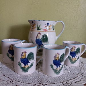 May include: Set of five white ceramic mugs with blue, green, and yellow rooster designs. The mugs have blue rims and handles. A matching white ceramic pitcher with a blue, green, and yellow rooster design is also included.