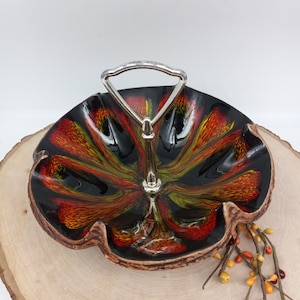 May include: A black ceramic serving dish with a wavy edge and a metal handle. The dish has a swirl pattern in shades of orange, yellow, and green.