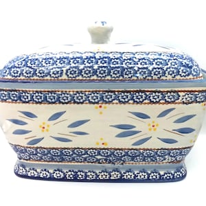 May include: A rectangular ceramic lidded casserole dish featuring a cream-colored base with hand-painted blue floral patterns, small yellow dots, and intricate blue and white geometric borders along the top and bottom edges of the vessel.