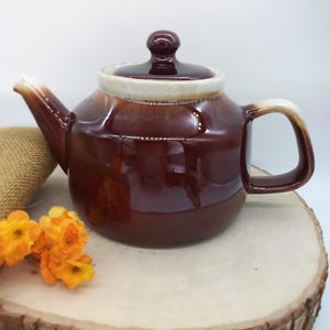 May include: A glossy dark brown ceramic teapot with a matching lid and a cream-colored rim. The teapot features a curved spout and a sturdy handle, both accented with cream-colored glaze at the top edges, presenting a smooth, reflective finish.
