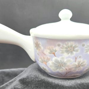 May include: A ceramic teapot with a side handle and a matching lid. The body of the teapot features a light purple background decorated with a pattern of white and grey floral blossoms. The handle and the lid are solid white in color.