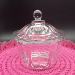 May include: A clear glass jar with a lid and a knob on top. The jar has a fluted design and is sitting on a pink crocheted mat.