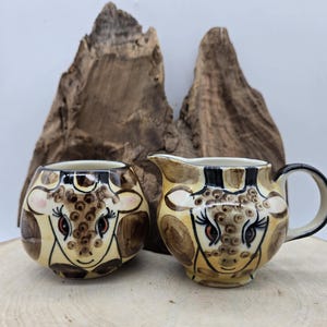 May include: A ceramic creamer and sugar bowl set. The set features a giraffe design with tan, brown, and black details. The creamer has a handle and spout, while the sugar bowl is round. The set is on a wooden surface.