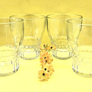 May include: Four clear drinking glasses with a textured base and a slightly flared top. A small arrangement of yellow and orange flowers is placed between the glasses. The glasses are empty.