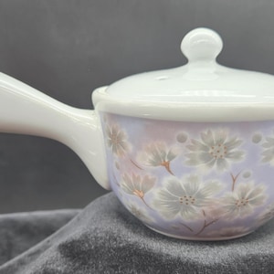 May include: A white ceramic serving bowl with a handle and lid. The bowl has a light purple base with a floral pattern in white and gray. The handle and lid are white.