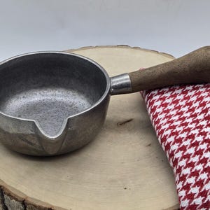 May include: A small, round, metallic pouring pan with a textured gray finish and a single integrated spout. The pan features a long, cylindrical wooden handle attached with a silver-toned metal ferrule, providing a sturdy grip for kitchen use.
