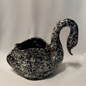May include: A ceramic swan-shaped planter with a speckled black and white pattern. The swan has a long, curved neck and a wide opening for planting. The planter has a textured surface and a glossy finish. The label on the swan reads "FLOOD WARE".
