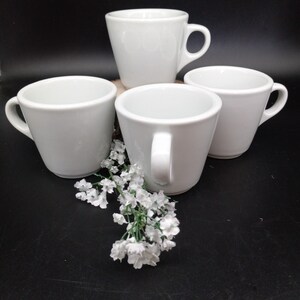 May include: Four white ceramic coffee cups with rounded handles. The cups are arranged in a circle on a dark surface with a sprig of white flowers in the center.