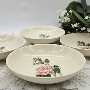 May include: Four off-white ceramic bowls with a floral rose design. Each bowl features a pink rose with green leaves. The bowls are arranged on a white lace tablecloth, with a blurred background of white flowers.