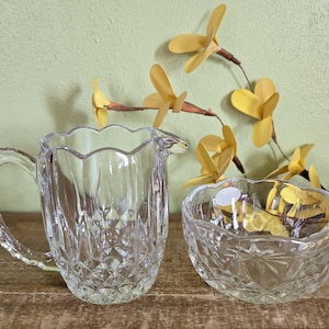 May include: A clear glass creamer and sugar bowl set with a cut glass design. The creamer has a handle and a scalloped rim. The sugar bowl is round with a scalloped rim.