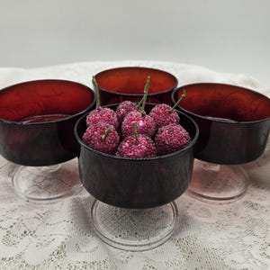 May include: Four dark red glass dessert bowls with clear glass stems. One bowl is filled with purple, berry-like objects. The bowls are arranged on a white lace tablecloth.