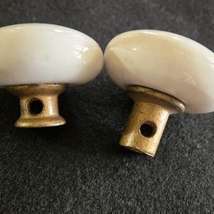 May include: Two vintage white ceramic door knobs with a gold-colored base. The knobs have an oval shape and are slightly off-white. The base has a hole for the screw. These knobs are suitable for furniture or doors.