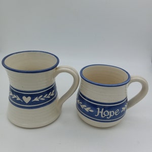 May include: Two ceramic mugs with blue and white designs. One mug has a heart and leaves design, the other has the word "Hope" with leaves.