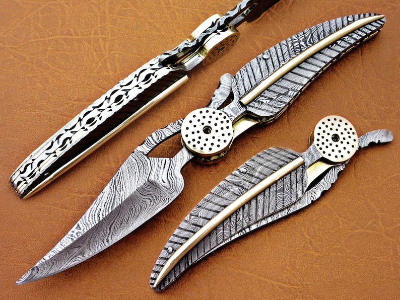 May include: Three folding pocket knives with intricate patterned blades and brass accents. The blades are made of Damascus steel and have a feather-like design.