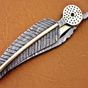 May include: A silver and gold pocket knife with a feather-shaped blade and a decorative brass cap. The blade is made of Damascus steel and has a unique pattern.