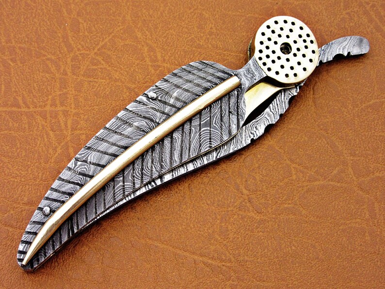 May include: A silver and gold pocket knife with a feather-shaped blade and a decorative brass cap. The blade is made of Damascus steel and has a unique pattern.