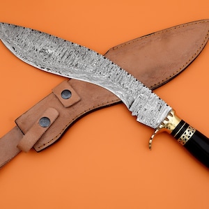 Może przedstawiać: Zakrzywiony nóż w kolorze srebrnym z czarną rękojeścią i złotym akcentem. Nóż znajduje się w brązowej skórzanej pochwie z paskiem.