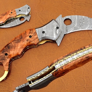 May include: A folding karambit knife with a Damascus steel blade and a wood handle. The handle has a brass ring on the end and a decorative pattern on the side.