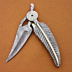 May include: A silver and gold pocket knife with a feather-shaped blade and a patterned handle. The blade is open and the knife is lying on a brown leather surface.