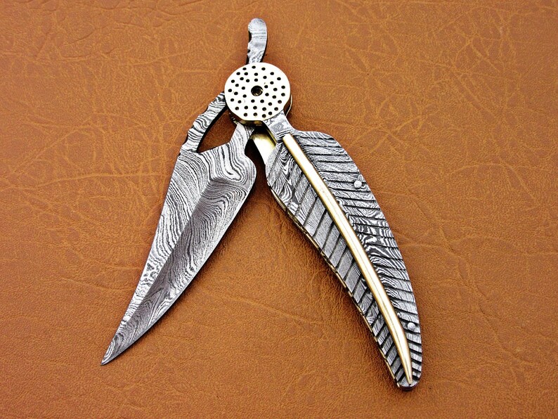 May include: A silver and gold pocket knife with a feather-shaped blade and a patterned handle. The blade is open and the knife is lying on a brown leather surface.