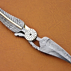 May include: A silver and gold pocket knife with a feather-shaped design on the handle. The blade is made of Damascus steel and has a wavy pattern.