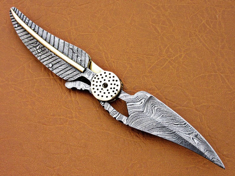 May include: A silver and gold pocket knife with a feather-shaped design on the handle. The blade is made of Damascus steel and has a wavy pattern.