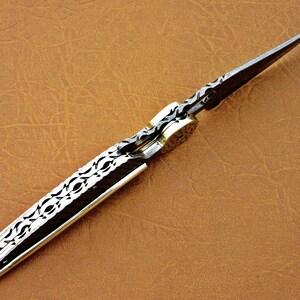 May include: A silver pocket knife with a patterned blade and a brass handle. The blade is open and the knife is lying on a brown leather surface.