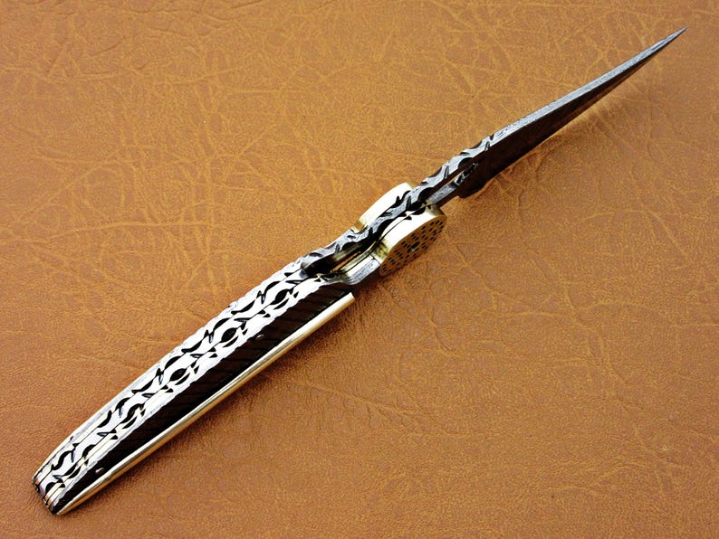 May include: A silver pocket knife with a patterned blade and a brass handle. The blade is open and the knife is lying on a brown leather surface.