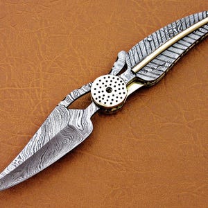 May include: A silver and gold pocket knife with a patterned blade and a feather-shaped handle.