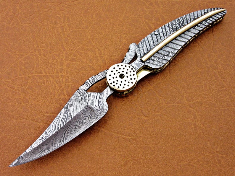 May include: A silver and gold pocket knife with a patterned blade and a feather-shaped handle.