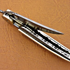May include: A silver pocket knife with a patterned blade and a white handle. The blade is open and the knife is lying on a brown leather surface.