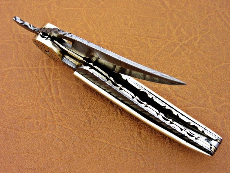May include: A silver pocket knife with a patterned blade and a white handle. The blade is open and the knife is lying on a brown leather surface.