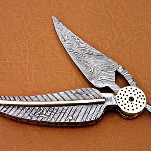 May include: A silver pocket knife with a patterned blade and a brass handle. The blade is open and the handle is shaped like a feather.
