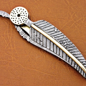 May include: A silver and gold pocket knife with a feather-shaped blade and a decorative brass cap. The blade has a damascus pattern.