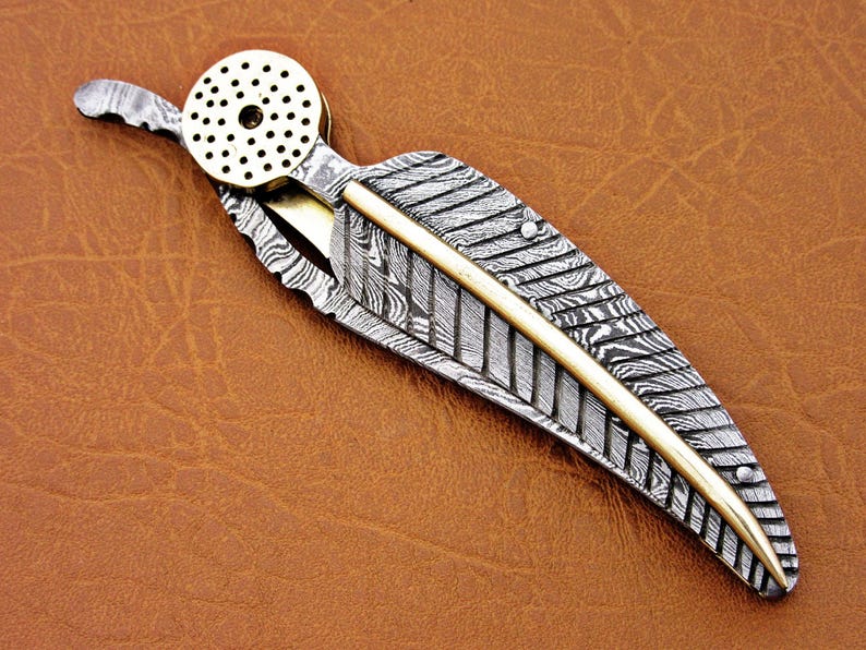 May include: A silver and gold pocket knife with a feather-shaped blade and a decorative brass cap. The blade has a damascus pattern.