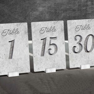 Puede incluir: Primer plano de tres tarjetas rectangulares con números de mesa. Cada tarjeta gris tiene la palabra "Table" en fuente cursiva. Los números 1, 15 y 30 están tallados en las tarjetas. Cada tarjeta está soportada por un pequeño soporte blanco.