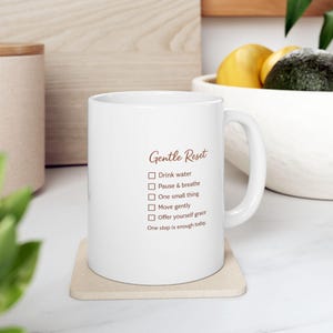 May include: White ceramic mug with the text "Gentle Reset" in a script font. The mug features a checklist with prompts like "Drink water" and "Pause & breathe." The mug sits on a beige coaster.