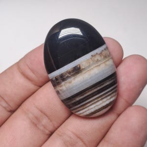 May include: An oval-shaped polished gemstone with a black top and layered bands of white, tan, and brown. The stone is held in a hand, showcasing its smooth, reflective surface. The gemstone is a natural, earthy product.