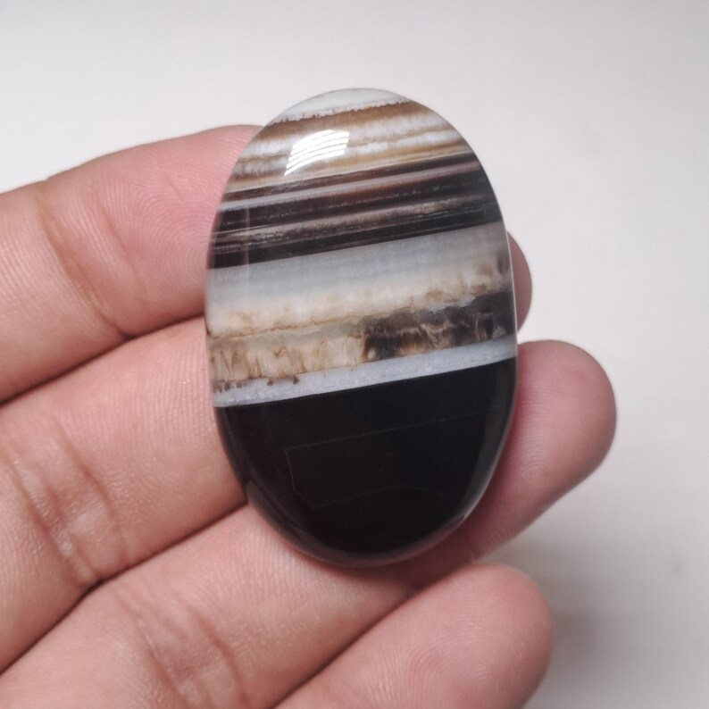 May include: An oval-shaped gemstone with a polished surface. The stone features layers of black, brown, white, and tan hues. The gemstone is held in a hand, showcasing its size and shape. The stone's design is natural and organic.