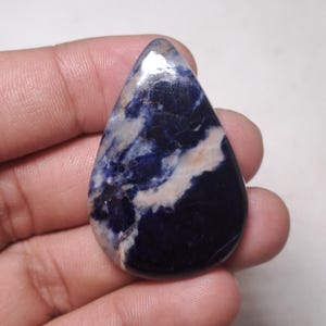 May include: A polished, teardrop-shaped sodalite gemstone. The stone features a deep blue base with swirling white and beige patterns. The surface is smooth and reflective, showcasing the natural variations in color and texture. This gemstone is ideal for jewelry making.